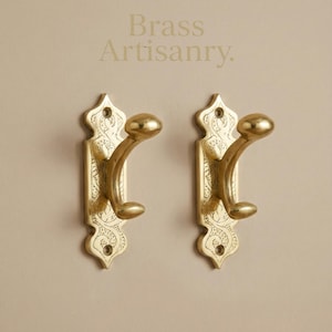 May include: Two brass artisanry wall hooks with decorative scrollwork and a curved hook design. The hooks are mounted on a neutral background, showcasing their ornate details and warm, metallic finish.