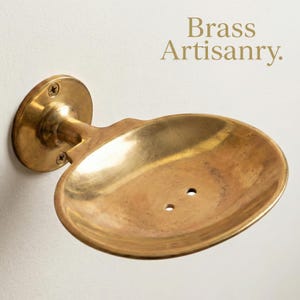 May include: A wall-mounted brass soap dish. The oval dish has two drainage holes and is connected to a circular wall plate via a curved arm. The text "Brass Artisanry" is visible. The fixture is attached to a white wall.
