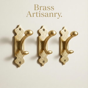 May include: Three brass wall hooks with an ornate, engraved design. The hooks have a curved shape with a rounded knob at the end. The text "Brass Artisanry" is above the hooks.
