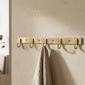 Op de afbeelding: Een messing wandhaak met zes gebogen haken, gemonteerd aan een muur. De rechthoekige haak heeft een getextureerd oppervlak. Een lichtgekleurde handdoek hangt aan een van de haken. De tekst "Brass Artisanry" is zichtbaar.
