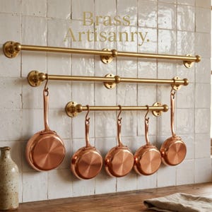 May include: A kitchen scene featuring three brass pot racks mounted on a white tiled wall. Five copper saucepans hang from the middle rack. The text "Brass Artisanry" is in the upper right corner.