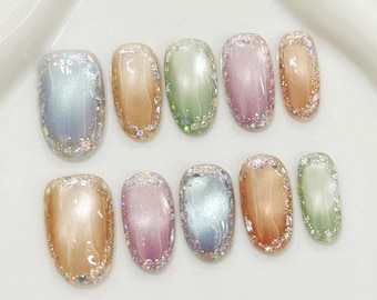 May include: A set of ten oval-shaped artificial nails. Each nail features a different pastel color, including blue, orange, green, pink, and peach. The edges of each nail are adorned with iridescent glitter, creating a shimmering effect.