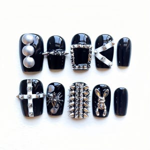 Puede incluir: Conjunto de diez uñas postizas negras con detalles plateados y blancos. Las uñas presentan varios diseños, incluyendo pinchos, una cruz, una araña, un conejo y un marco cuadrado.