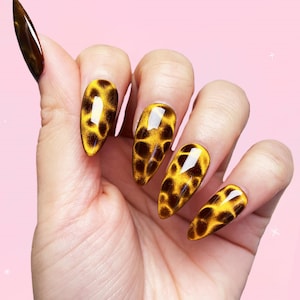 May include: A hand displaying long, almond-shaped artificial nails. The nails feature a yellow base with a dark brown leopard print design. The thumb nail is a solid dark brown color.