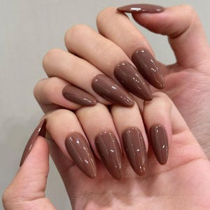 May include: A hand displaying long, almond-shaped, brown artificial nails. The nails have a glossy finish and are a solid, warm brown color. The nails are well-manicured and evenly shaped.