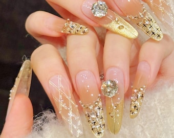 May include: Close-up of long, embellished fingernails. The nails feature a gradient design, transitioning from a natural pink to a gold hue. They are adorned with sparkling rhinestones, gold accents, and intricate patterns, creating a glamorous aesthetic.