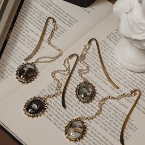 May include: Ornate gold-toned bookmarks with oval pendants featuring miniature landscape and bird art, connected by delicate chains. The bookmarks rest on the pages of an open book, creating a vintage aesthetic.