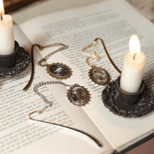May include: An open book with three antique-style pendant necklaces and a metal bookmark. Two lit candles in ornate holders flank the book. The necklaces have oval pendants with miniature scenes and hang on delicate chains.