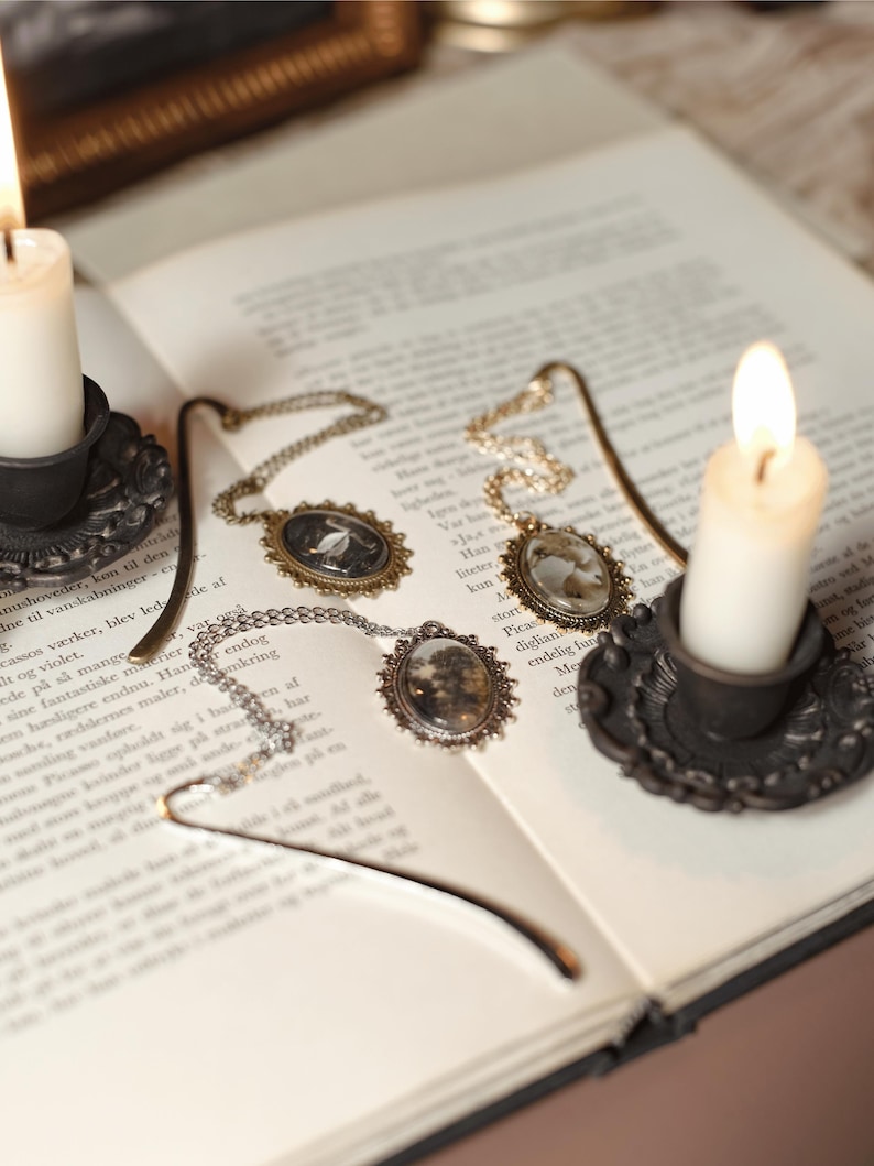 May include: An open book with three antique-style pendant necklaces and a metal bookmark. Two lit candles in ornate holders flank the book. The necklaces have oval pendants with miniature scenes and hang on delicate chains.
