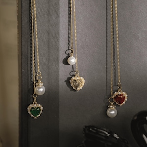 May include: Gold-toned necklaces with heart-shaped pendants and pearl accents. One pendant is emerald green, another is gold, and the third is ruby red. The necklaces hang against a dark gray background.