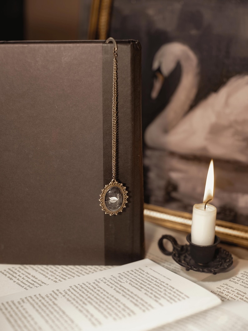 May include: A dark brown book with a decorative gold-colored bookmark featuring a small oval pendant with a swan design. A lit white candle in a black holder sits nearby, with a swan painting in the background.