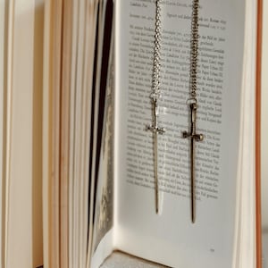 May include: Two silver-toned sword-shaped bookmarks hanging from the top of an open book. The book's pages are aged, with German text visible. The bookmarks are suspended by chains.