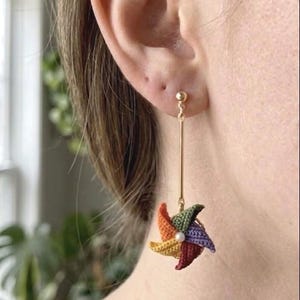 May include: A gold-toned drop earring featuring a small, multi-colored crocheted pinwheel charm. The pinwheel is composed of orange, yellow, green, purple, and red thread segments, centered with a tiny white bead, hanging from a thin gold bar.