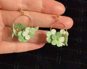 May include: A pair of gold-colored hoop earrings adorned with delicate, light green and white crocheted flowers. The earrings feature a small gold stud at the top and are set against a dark background.