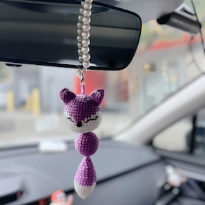 Op de afbeelding: Paarse gehaakte vos autohanger die hangt aan een witte parelketting. De hanger is bevestigd aan de achteruitkijkspiegel van een auto.