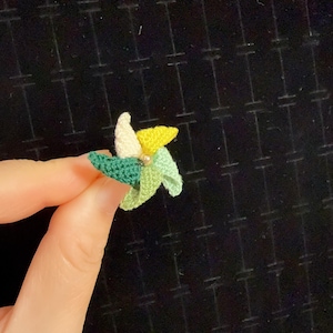 Puede incluir: Un anillo de ganchillo hecho a mano con forma de flor. El anillo presenta pétalos en tonos verdes, amarillos y blancos, con una pequeña cuenta en el centro. El anillo es sostenido por una mano sobre un fondo negro.