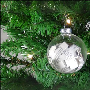 May include: A clear glass Christmas ornament filled with crumpled pieces of white paper with black text. The ornament is hanging on a green artificial Christmas tree.