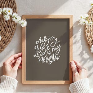 Pode incluir: Uma impress&atilde;o emoldurada com o texto "the joy of the Lord is my strength" em escrita branca. A impress&atilde;o tem um fundo cinza escuro e uma moldura de madeira clara. A imagem &eacute; cercada por esteiras tecidas e flores brancas.