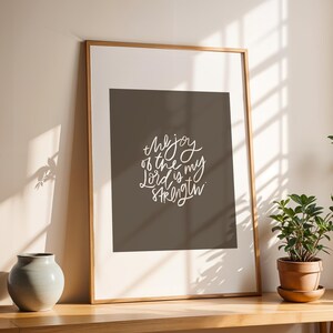 Pode incluir: Uma impress&atilde;o emoldurada com o texto "the joy of the Lord is my strength" em escrita branca. A impress&atilde;o &eacute; exibida em uma mesa de madeira, ao lado de uma pequena planta em vaso e um vaso cinza. A moldura &eacute; de cor marrom claro.