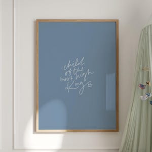 Könnte beinhalten: Ein rechteckiges Wandbild in einem hellen Holzrahmen mit einem staubblauen Hintergrund, das den weißen kursiven Schriftzug "child of the most high King" und ein kleines Kronensymbol zeigt, platziert über einem hölzernen Kinderbett.