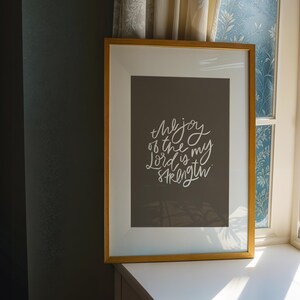 Pode incluir: Uma impress&atilde;o emoldurada com uma moldura dourada. A impress&atilde;o tem um fundo cinza escuro com texto branco em it&aacute;lico que diz "the joy of the Lord is my strength". A impress&atilde;o emoldurada est&aacute; em uma superf&iacute;cie branca perto de uma janela.
