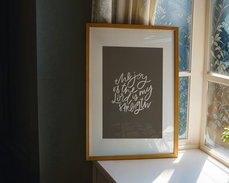 Pode incluir: Uma impress&atilde;o emoldurada com uma moldura dourada. A impress&atilde;o tem um fundo cinza escuro com texto branco em it&aacute;lico que diz "the joy of the Lord is my strength". A impress&atilde;o emoldurada est&aacute; em uma superf&iacute;cie branca perto de uma janela.