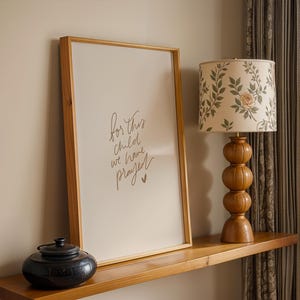 Könnte beinhalten: Ein holzgerahmter Druck mit dem Text "for this child we have prayed" steht auf einem Holzregal. Eine floral gemusterte Lampe mit Holzfuß und ein schwarzer Keramiktopf befinden sich ebenfalls auf dem Regal.