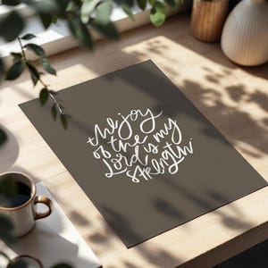 Pode incluir: Uma impress&atilde;o com o texto "the joy of the Lord is my strength" em escrita branca sobre um fundo cinza escuro. A impress&atilde;o est&aacute; sobre uma superf&iacute;cie de madeira, com uma caneca de caf&eacute; e vegeta&ccedil;&atilde;o no fundo.