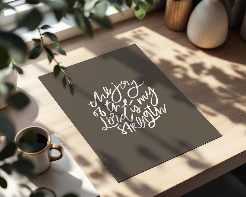 Pode incluir: Uma impress&atilde;o com o texto "the joy of the Lord is my strength" em escrita branca sobre um fundo cinza escuro. A impress&atilde;o est&aacute; sobre uma superf&iacute;cie de madeira, com uma caneca de caf&eacute; e vegeta&ccedil;&atilde;o no fundo.