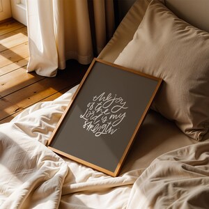 Pode incluir: Uma impress&atilde;o emoldurada com o texto "The joy of the Lord is my strength" em escrita branca sobre um fundo cinza escuro. A moldura de madeira repousa sobre uma cama com roupa de cama e almofadas bege. A cena &eacute; iluminada por luz natural.