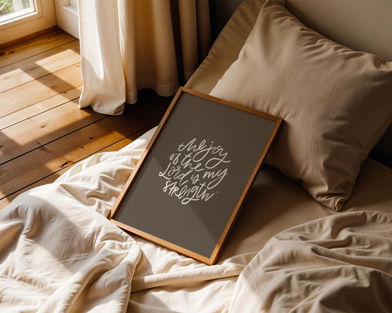 Pode incluir: Uma impress&atilde;o emoldurada com o texto "The joy of the Lord is my strength" em escrita branca sobre um fundo cinza escuro. A moldura de madeira repousa sobre uma cama com roupa de cama e almofadas bege. A cena &eacute; iluminada por luz natural.