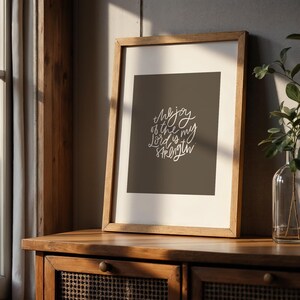 Pode incluir: Uma impress&atilde;o emoldurada com o texto "The joy of the Lord is my strength" em escrita branca sobre um fundo cinza escuro. A moldura de madeira est&aacute; sobre um arm&aacute;rio de madeira, com uma janela e vegeta&ccedil;&atilde;o ao fundo.