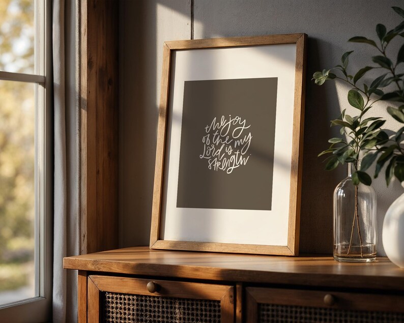 Pode incluir: Uma impress&atilde;o emoldurada com o texto "The joy of the Lord is my strength" em escrita branca sobre um fundo cinza escuro. A moldura de madeira est&aacute; sobre um arm&aacute;rio de madeira, com uma janela e vegeta&ccedil;&atilde;o ao fundo.