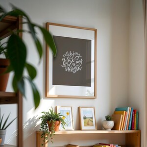 Pode incluir: Uma impress&atilde;o emoldurada com o texto "the joy of the Lord is my strength" em escrita branca. A impress&atilde;o &eacute; exibida em uma parede acima de uma prateleira de madeira com plantas em vasos e obras de arte emolduradas. Uma variedade de livros coloridos tamb&eacute;m est&atilde;o na prateleira.