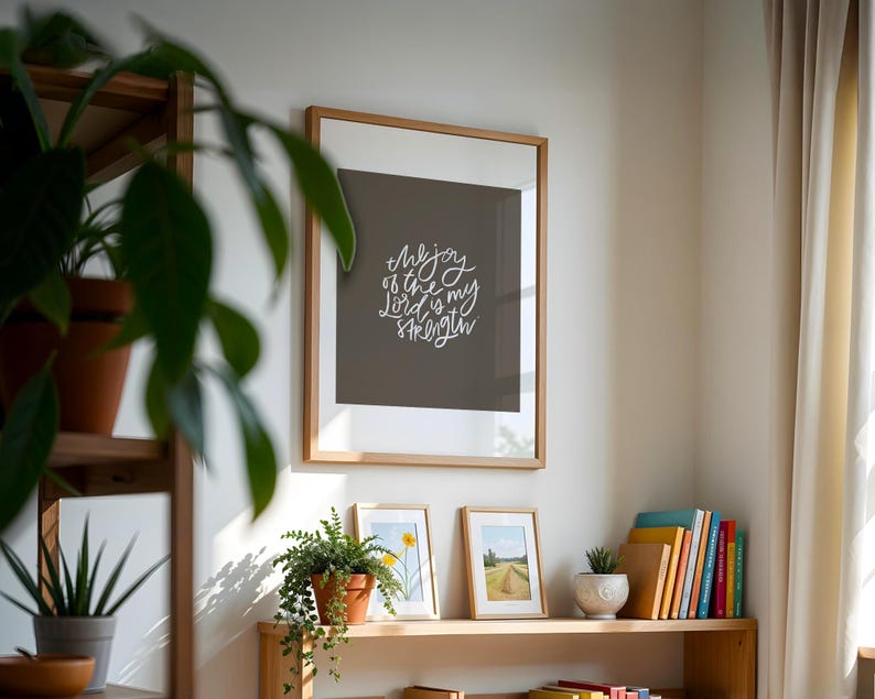 Pode incluir: Uma impress&atilde;o emoldurada com o texto "the joy of the Lord is my strength" em escrita branca. A impress&atilde;o &eacute; exibida em uma parede acima de uma prateleira de madeira com plantas em vasos e obras de arte emolduradas. Uma variedade de livros coloridos tamb&eacute;m est&atilde;o na prateleira.