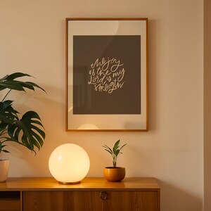 Pode incluir: Uma impress&atilde;o emoldurada com o texto "the joy of the Lord is my strength" est&aacute; pendurada acima de um arm&aacute;rio de madeira. Uma l&acirc;mpada redonda e brilhante est&aacute; sobre o arm&aacute;rio, ao lado de uma planta em vaso. Uma planta grande e frondosa est&aacute; em um vaso branco.