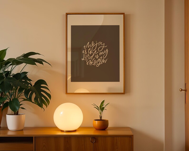 Pode incluir: Uma impress&atilde;o emoldurada com o texto "the joy of the Lord is my strength" est&aacute; pendurada acima de um arm&aacute;rio de madeira. Uma l&acirc;mpada redonda e brilhante est&aacute; sobre o arm&aacute;rio, ao lado de uma planta em vaso. Uma planta grande e frondosa est&aacute; em um vaso branco.