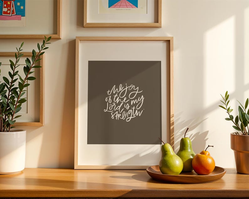 Pode incluir: Uma impress&atilde;o emoldurada com o texto "the joy of the Lord is my strength" em escrita branca. Duas peras verdes e uma fruta laranja est&atilde;o em um prato de madeira. Uma planta em vaso est&aacute; &agrave; direita.