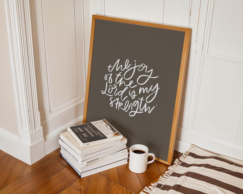 Pode incluir: Uma impress&atilde;o emoldurada com o texto "the joy of the Lord is my strength" em escrita branca. A impress&atilde;o est&aacute; encostada a uma parede ao lado de uma pilha de livros, uma caneca de caf&eacute; branca e um tapete listrado.