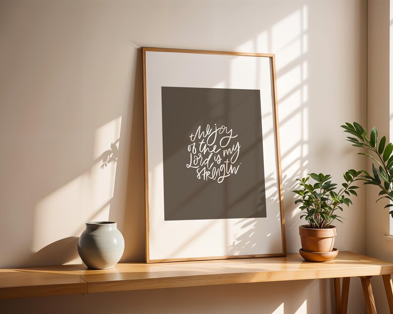 Pode incluir: Uma impress&atilde;o emoldurada com o texto "the joy of the Lord is my strength" em escrita branca. A impress&atilde;o &eacute; exibida em uma mesa de madeira, ao lado de uma pequena planta em vaso e um vaso cinza. A moldura &eacute; de cor marrom claro.