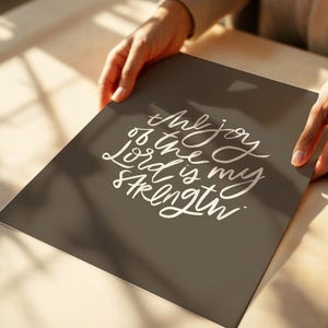 Pode incluir: Uma impress&atilde;o retangular com o texto "the joy of the Lord is my strength" em escrita branca sobre um fundo cinza escuro. A impress&atilde;o &eacute; segurada por duas m&atilde;os, com uma superf&iacute;cie de madeira ao fundo.