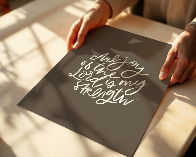 Pode incluir: Uma impress&atilde;o retangular com o texto "the joy of the Lord is my strength" em escrita branca sobre um fundo cinza escuro. A impress&atilde;o &eacute; segurada por duas m&atilde;os, com uma superf&iacute;cie de madeira ao fundo.
