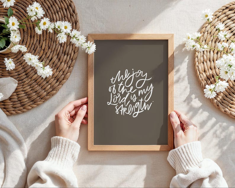 Pode incluir: Uma impress&atilde;o emoldurada com o texto "the joy of the Lord is my strength" em escrita branca. A impress&atilde;o tem um fundo cinza escuro e uma moldura de madeira clara. A imagem &eacute; cercada por esteiras tecidas e flores brancas.