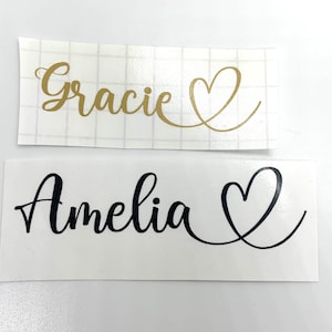 May include: Two vinyl decals featuring names and heart motifs. The top decal displays the name "Gracie" in gold script with a gold heart. The bottom decal shows the name "Amelia" in black script with a black heart.