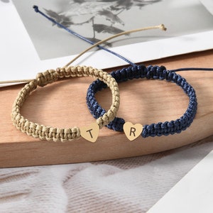 May include: Two macrame friendship bracelets. One is tan with a gold heart charm with the letter "T". The other is navy blue with a gold heart charm with the letter "R". Both bracelets have adjustable sliding knots.