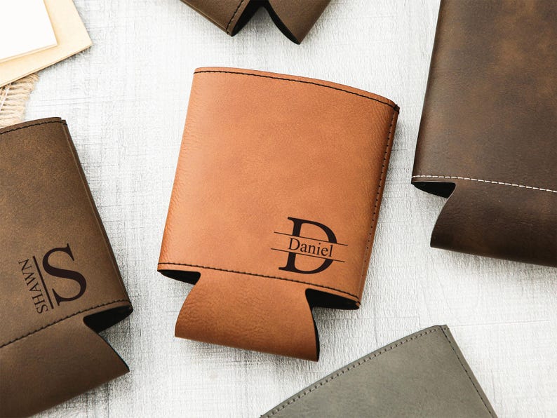 May include: A collection of personalized leatherette can coolers in various shades of brown and gray. One cooler is tan with the name "Daniel" and initial "D" engraved. Other coolers have initials and names.