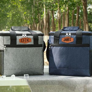 Pu&ograve; includere: Due borse frigo isolate, una grigia e una blu navy, con dettagli neri e toppe in pelle personalizzate. La borsa grigia ha "DK" e quella blu "JAMES". Entrambe hanno manici superiori e tasche laterali. Sono visibili bottiglie di birra e un bicchiere di birra.