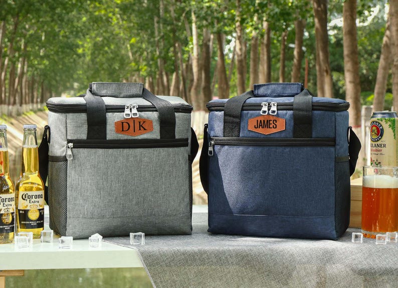 Pu&ograve; includere: Due borse frigo isolate, una grigia e una blu navy, con dettagli neri e toppe in pelle personalizzate. La borsa grigia ha "DK" e quella blu "JAMES". Entrambe hanno manici superiori e tasche laterali. Sono visibili bottiglie di birra e un bicchiere di birra.