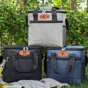 Pu&ograve; includere: Tre borse frigo personalizzate in grigio, nero e blu. Ogni borsa ha una toppa in similpelle marrone con iniziali. Le borse hanno manici e cerniere nere. Le borse sono su una coperta con bottiglie di birra.