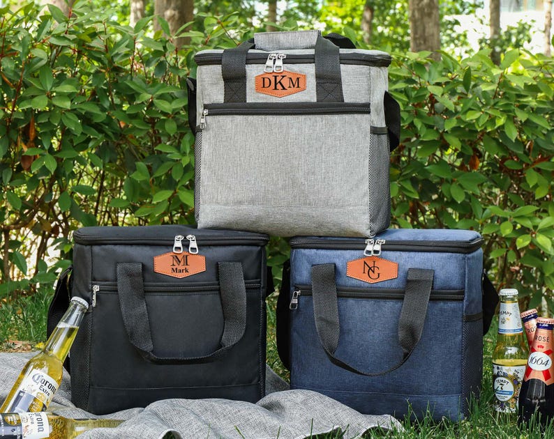 Pu&ograve; includere: Tre borse frigo personalizzate in grigio, nero e blu. Ogni borsa ha una toppa in similpelle marrone con iniziali. Le borse hanno manici e cerniere nere. Le borse sono su una coperta con bottiglie di birra.